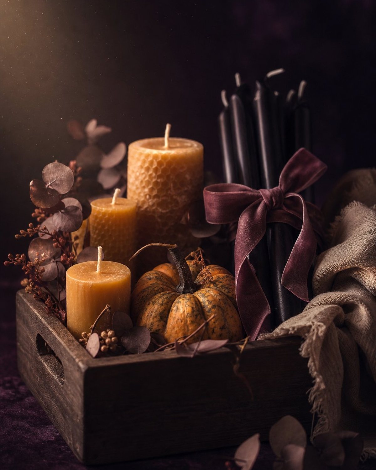 A walnut crate packed with amber beeswax candles, an heirloom pumpkin, dried eucalyptus and a bundle of black tapers tied with plum velvet ribbon.